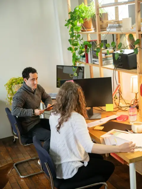 Un bărbat și o femeie lucrează împreună la un birou într-un studio plin de plante, bărbatul întinzând ceva peste masă.
