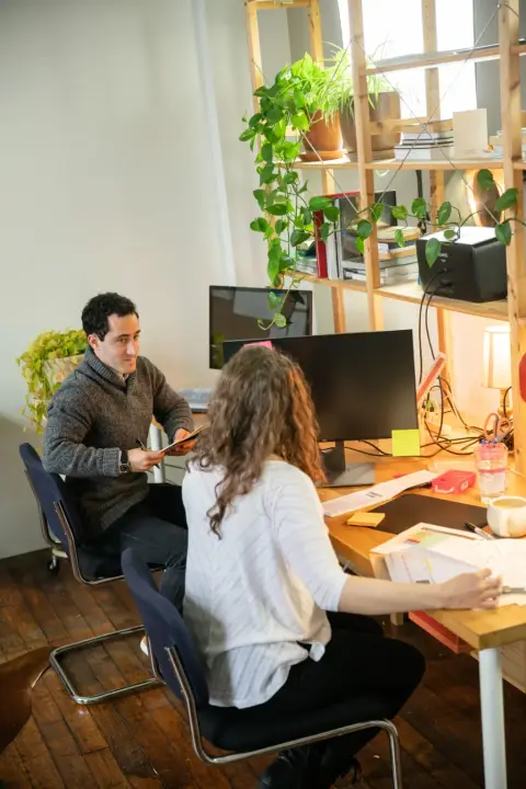 Un bărbat și o femeie lucrează împreună la un birou într-un studio plin de plante, bărbatul întinzând ceva peste masă.