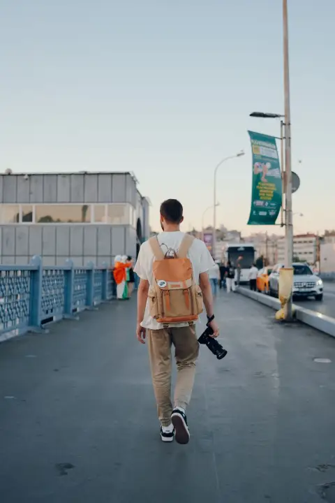 Un bărbat cu un rucsac și un aparat foto merge pe un pod din oraș în lumina apusului.