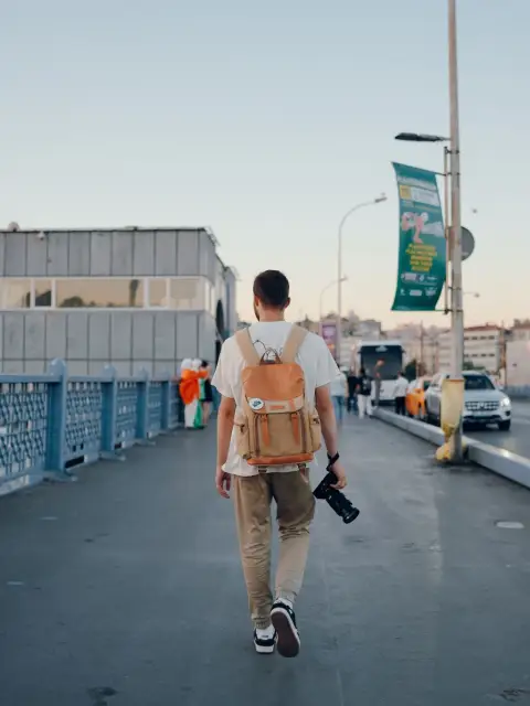 Un bărbat cu un rucsac și un aparat foto merge pe un pod din oraș în lumina apusului.