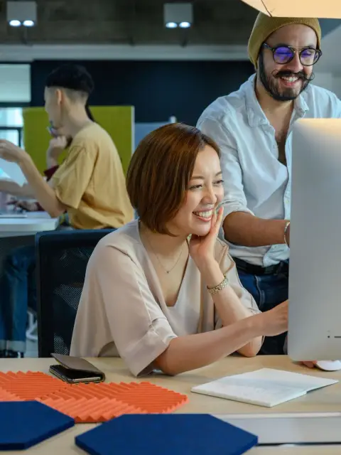 O femeie zâmbitoare lucrează la un monitor desktop într-un birou, în timp ce un coleg stă lângă ea.